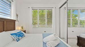 Un dormitorio con una cama, una almohada azul, una toalla enrollada y una ventana con vista a follaje.
