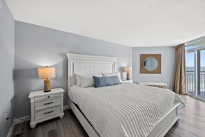 A modern bedroom with a large bed, white nightstands, a round mirror, and a view of the ocean through large windows. - Renovated Bay Watch 812 - 2 Bedroom/2 Bath (North Myrtle Beach)