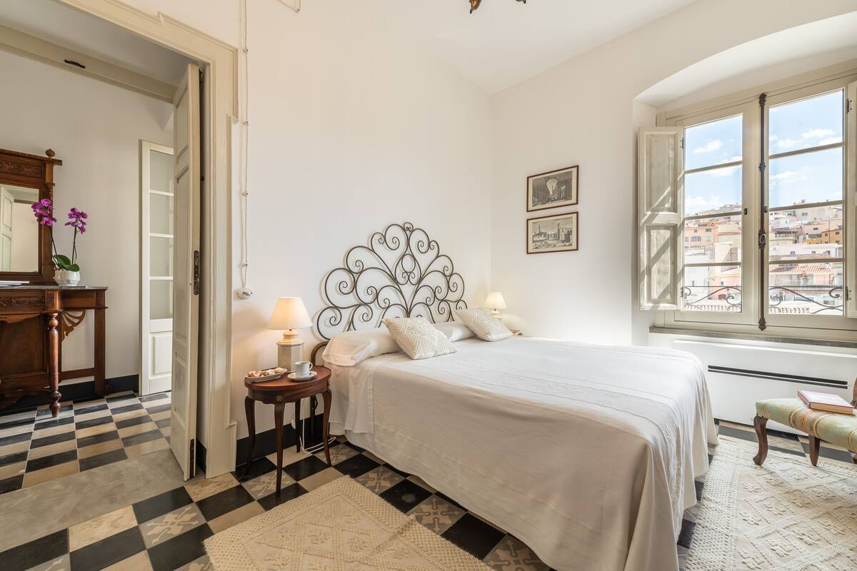 A bedroom with a checkered floor, a bed with white bedding, a nightstand with lamps, a mirror, and a window with a city view.