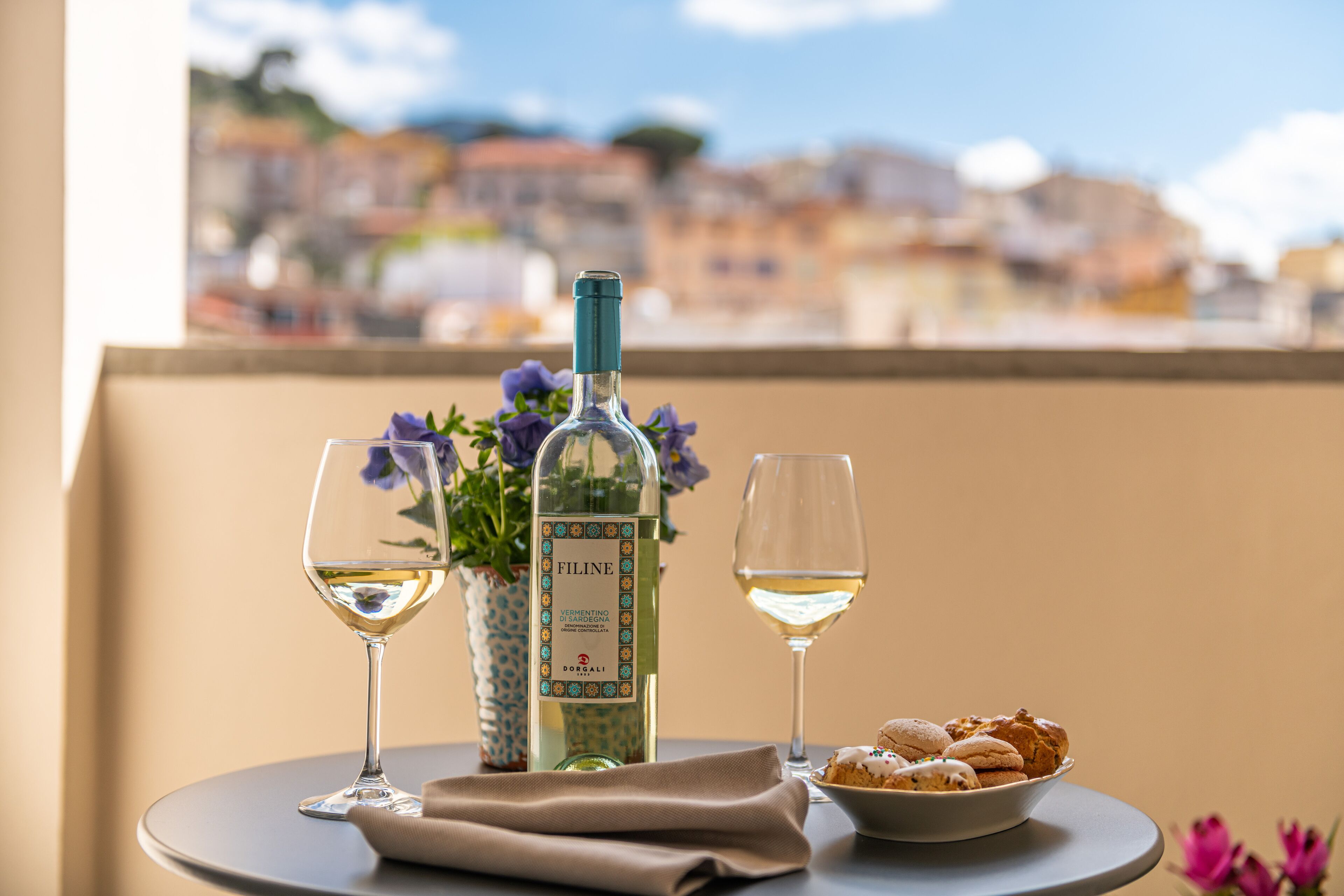 A table with a bottle of wine, two glasses, a bowl of cookies, and a vase of flowers.