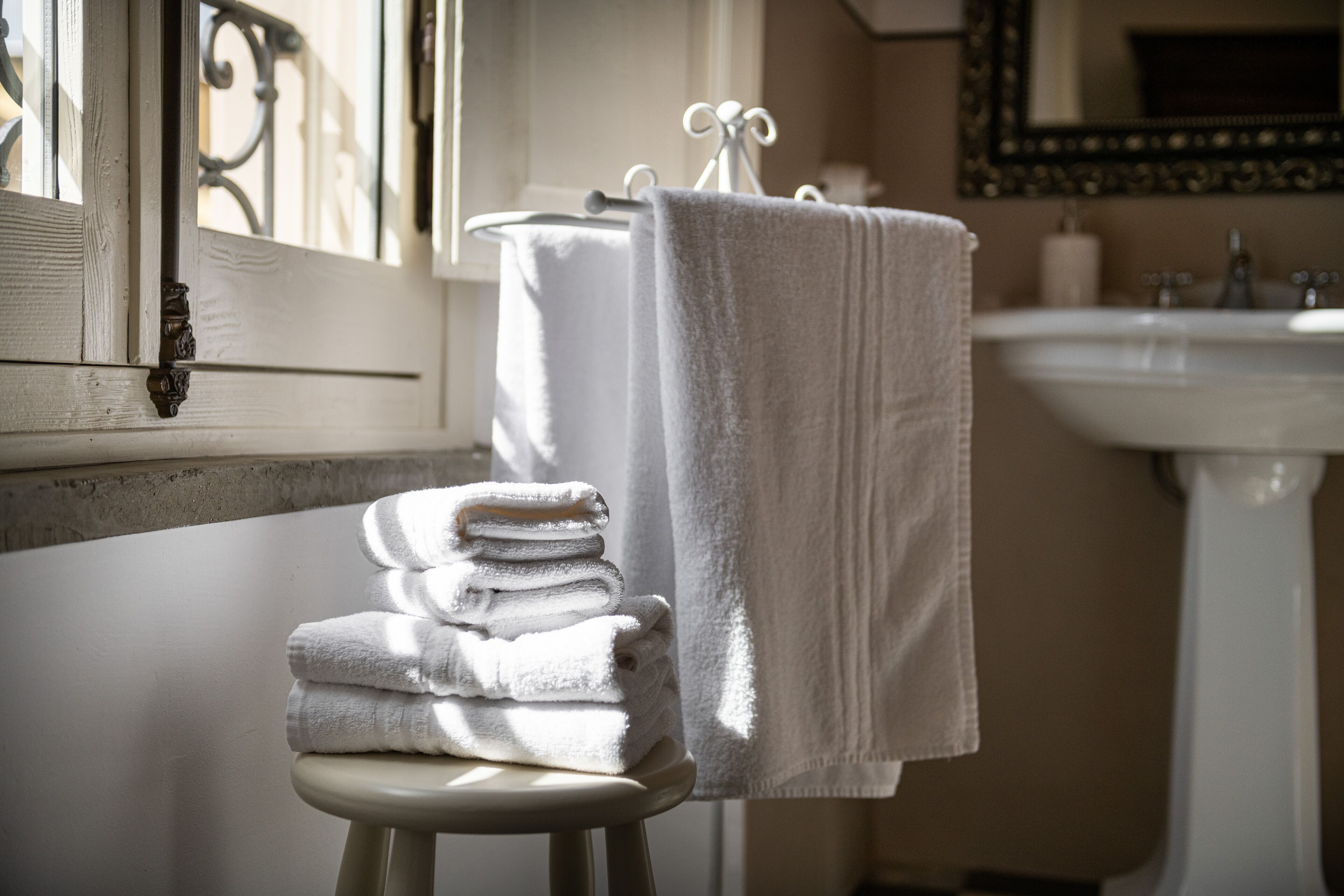 A bathroom with a stack of white towels on a stool, a white sink, and a window with decorative iron railings.
