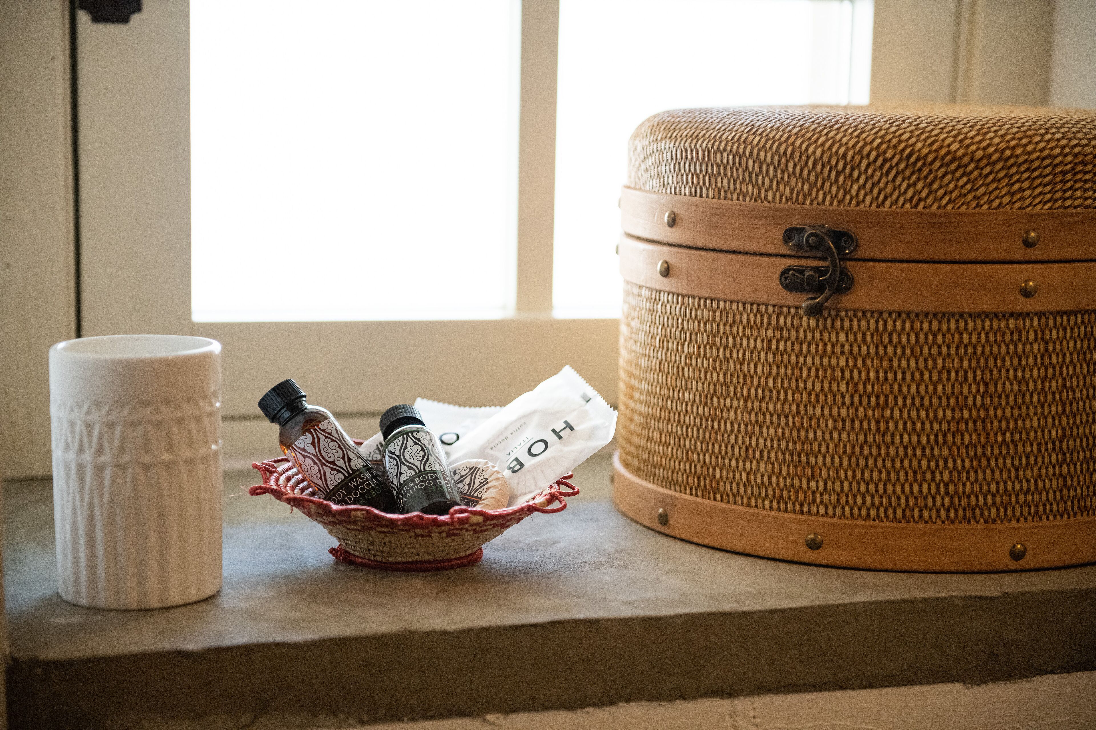 A wicker suitcase, a white ceramic mug, and a woven basket with toiletries on a windowsill.