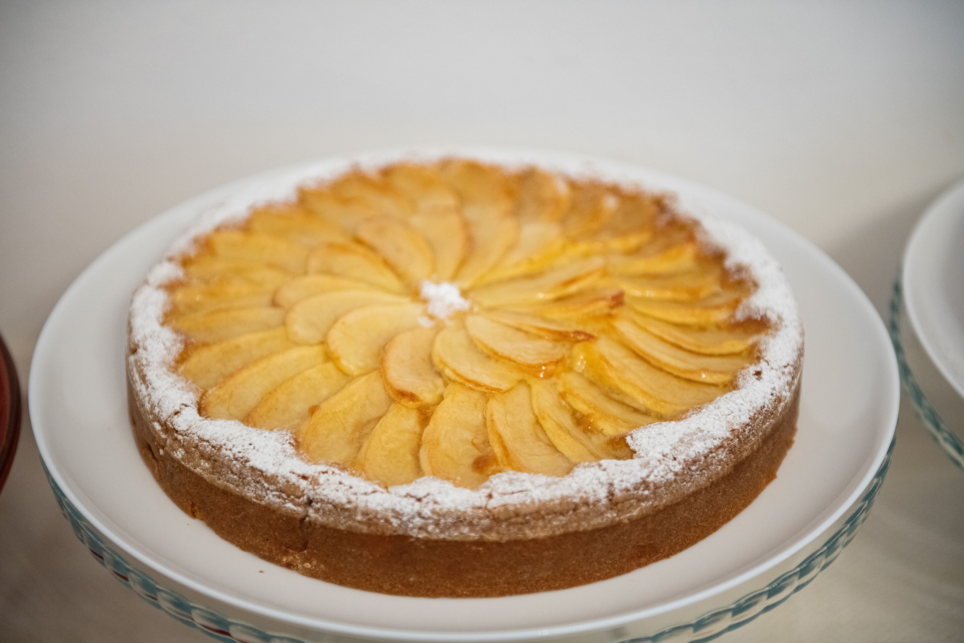 A round cake with sliced apples arranged in a circular pattern on top, dusted with powdered sugar, on a white plate.
