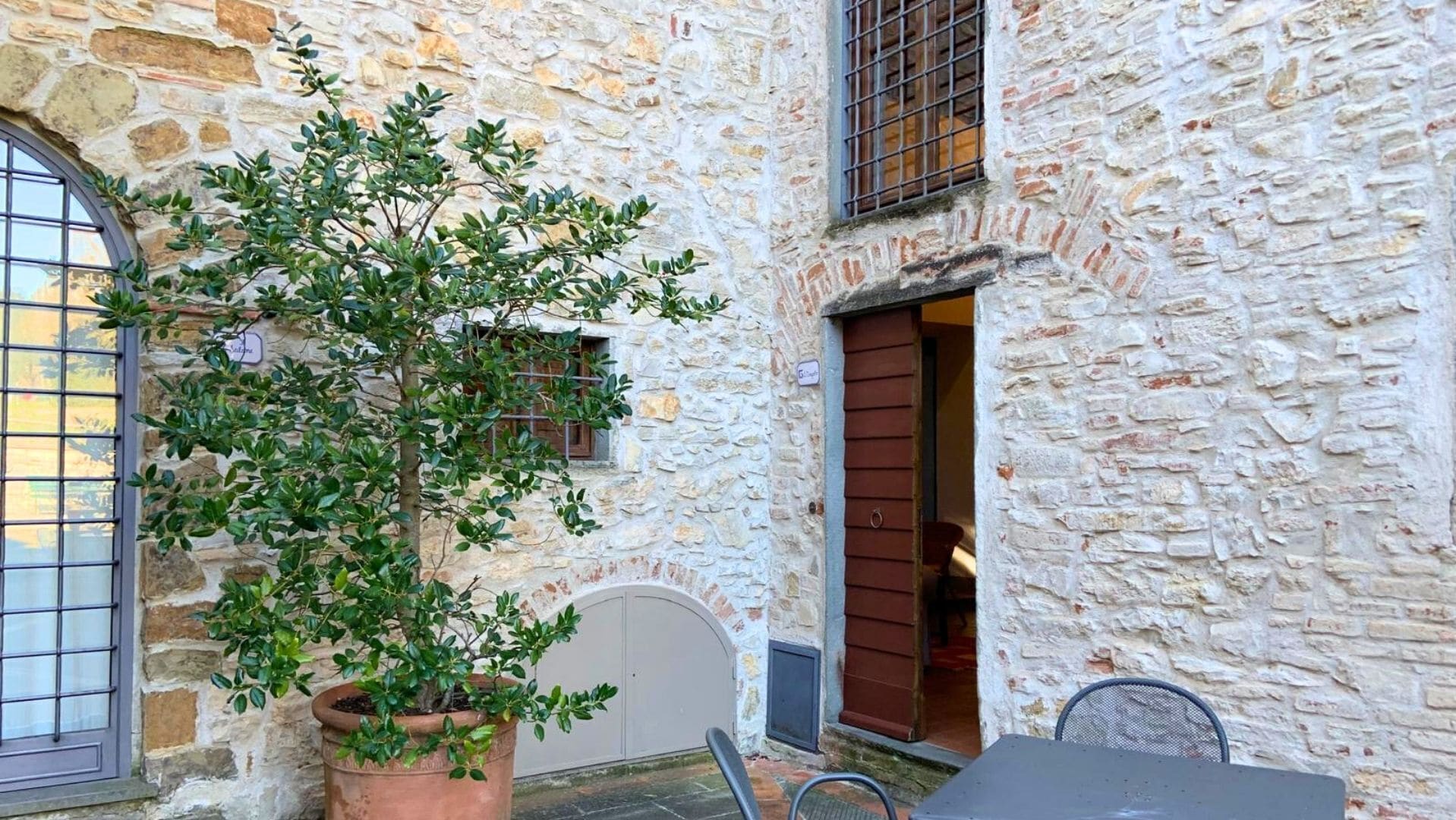 Edificio de piedra con un árbol en maceta al frente, una ventana con rejas y una puerta con un persiano marrón.