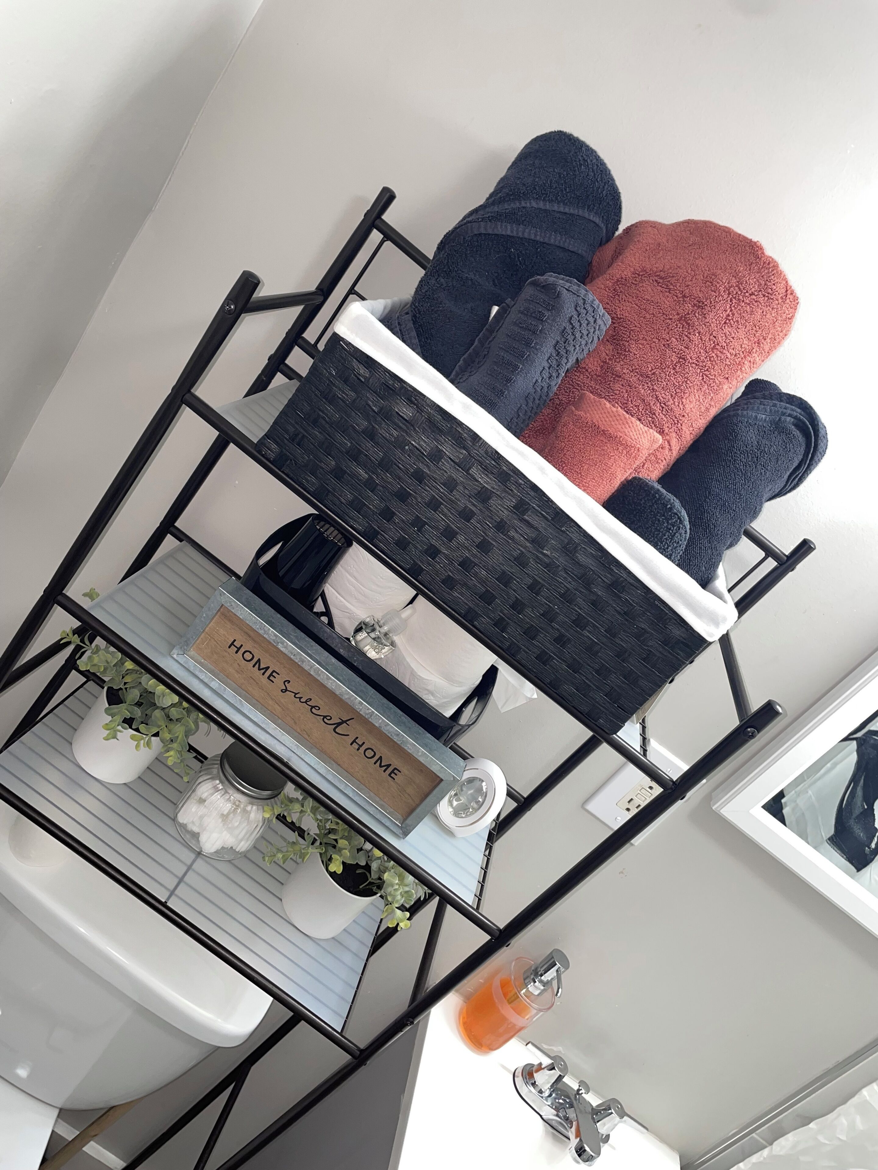 Ein Badezimmer mit einem schwarzen Metallregal, das Handtücher trägt, einer kleinen Ablage mit einem Topf und einem Schild mit der Aufschrift 'HOME SWEET HOME'.