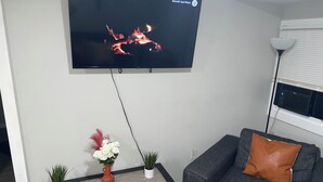 A living room with a flat-screen TV mounted on the wall, a grey sofa with a brown cushion, a small table with two potted plants, and a window with blinds.