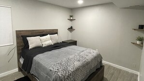 A bedroom with a bed, bedside tables, a nightstand, a wall-mounted shelf, and a potted plant.