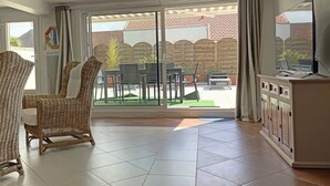 Un vaste espace de vie avec une grande porte-fenêtre coulissante donnant sur une terrasse extérieure. À l'intérieur, on trouve des fauteuils en osier, une table à manger et un buffet.