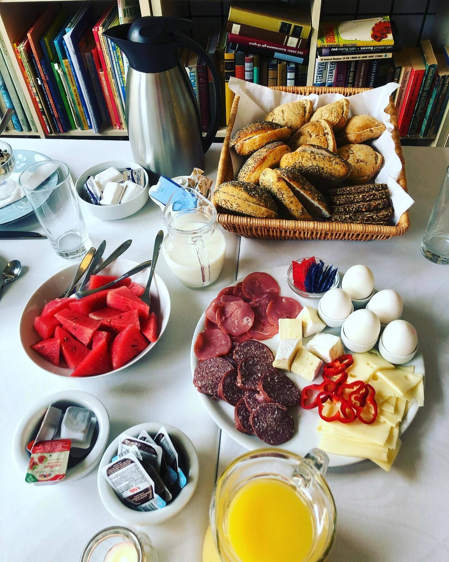 Una mesa de desayuno con diferentes alimentos, una tetera y una estantería al fondo.