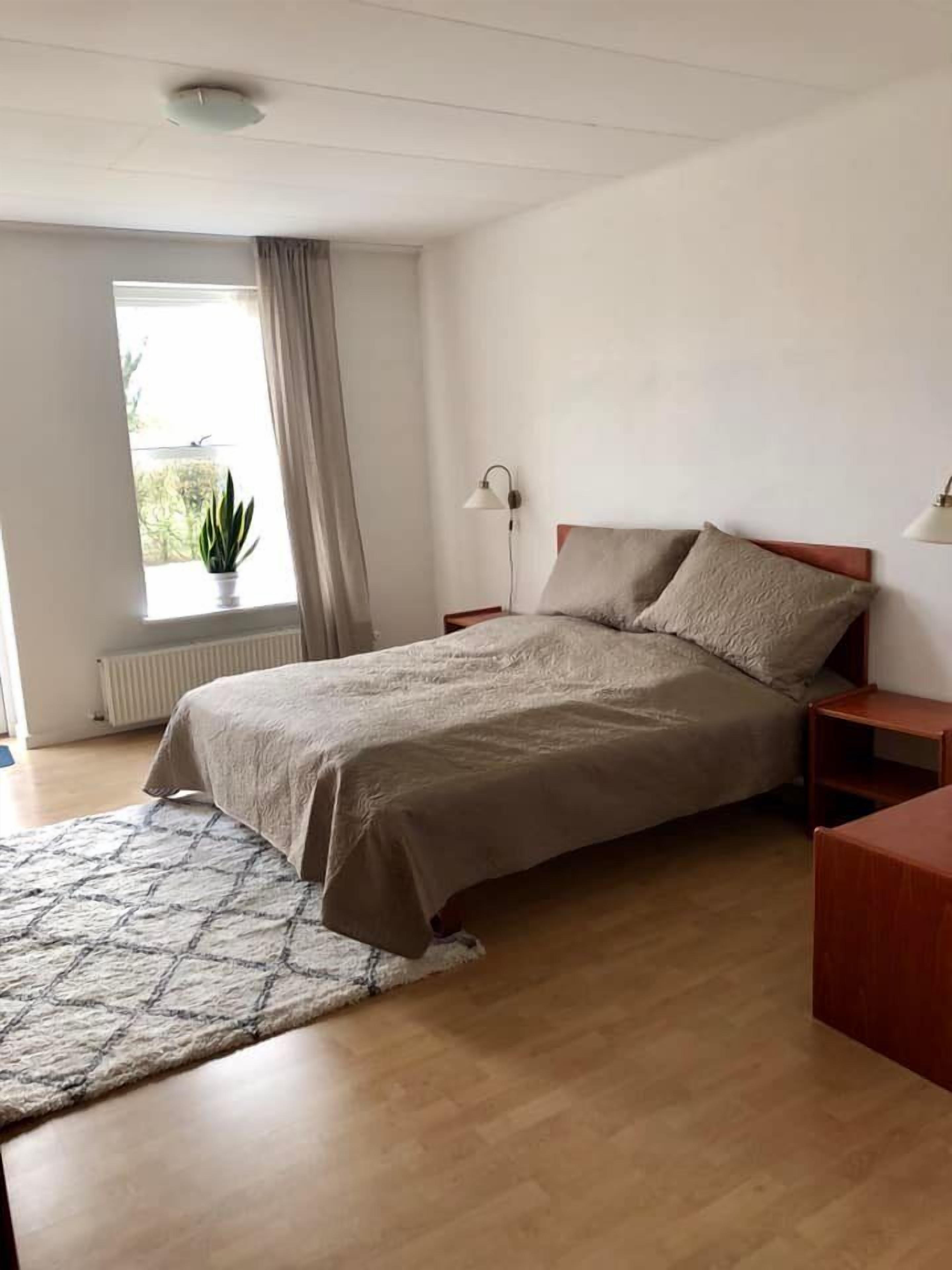 Un dormitorio con cama, mesita de noche, lámpara, ventana con cortinas y una planta en maceta.