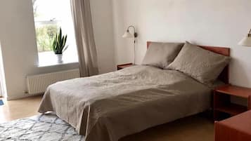 Un dormitorio con cama, mesita de noche, lámpara, ventana con cortinas y una planta en maceta.