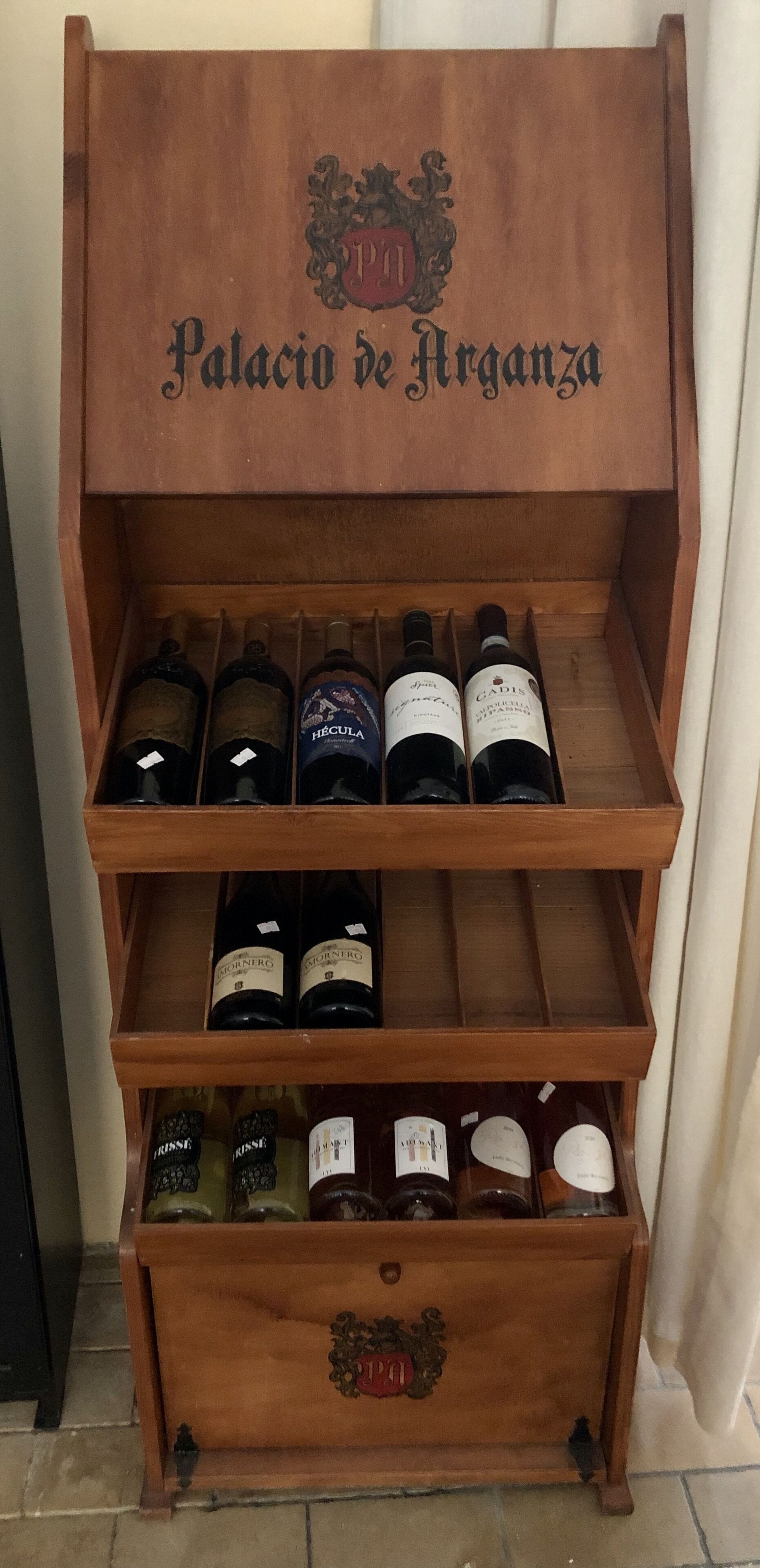 A wooden wine rack with multiple shelves, each holding bottles of wine. The top shelf has a label reading "Palacio de Arguenza."