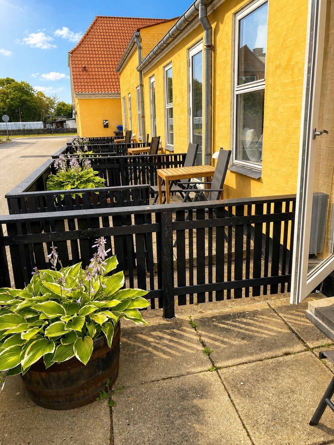 A yellow building with a red-tiled roof, outdoor seating with black chairs and tables, and a black fence.