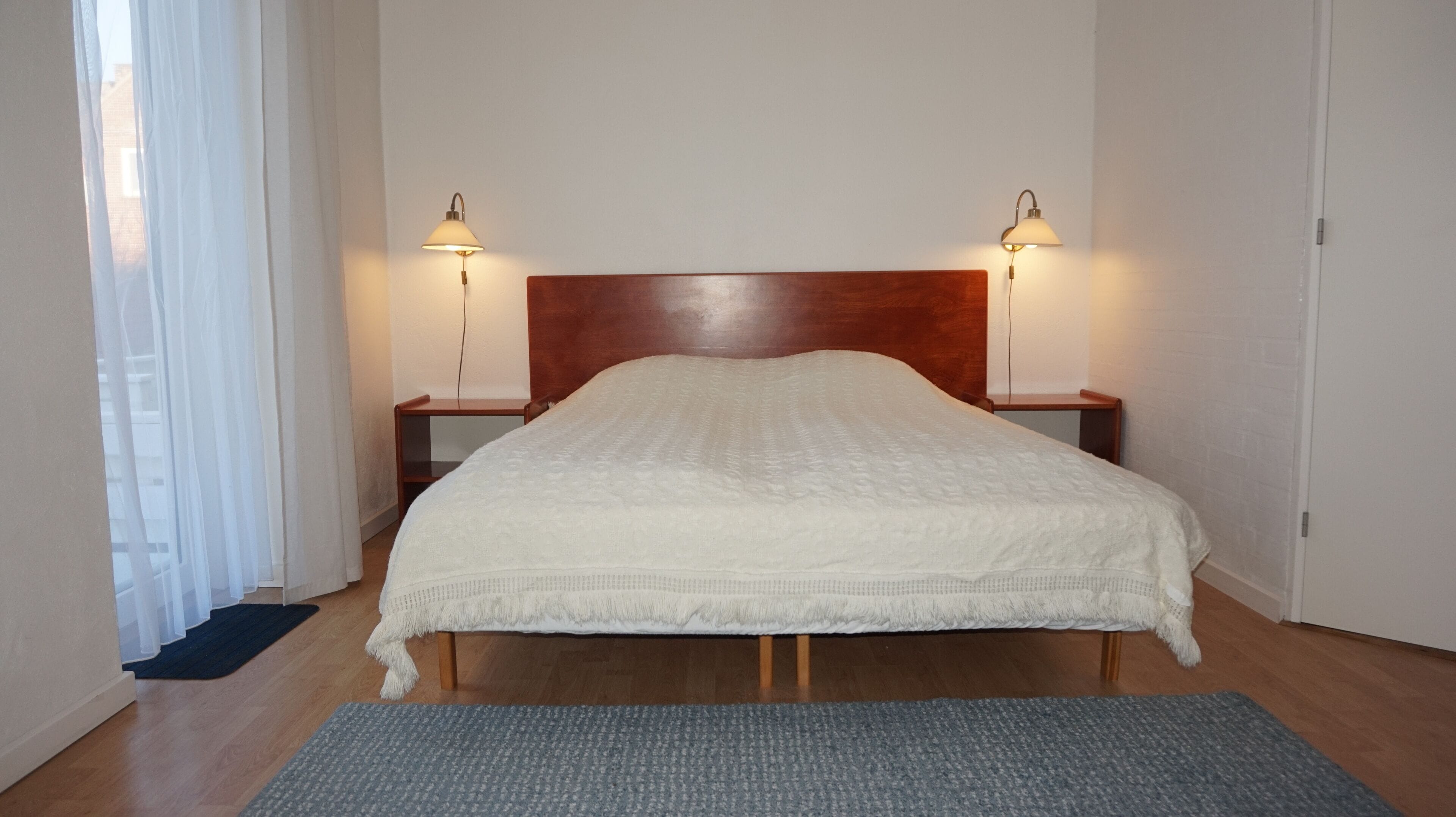 Uma cama bem arrumada com cabeceira de madeira, duas mesas de cabeceira com luminárias e um tapete cinza sobre o piso de madeira.