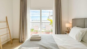 Une chambre d’hôtel avec un lit, une table de chevet avec une lampe, un balcon avec des chaises et une vue sur la mer.
