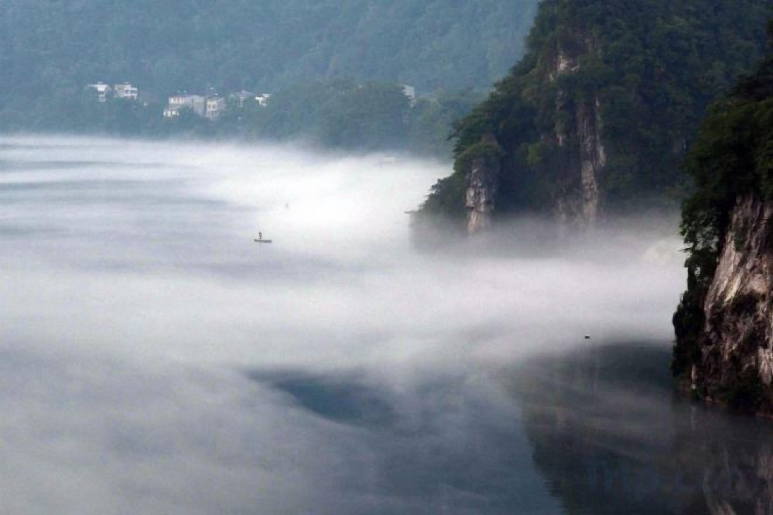 A river with a misty atmosphere, a cliff on one side, and a small boat in the distance.