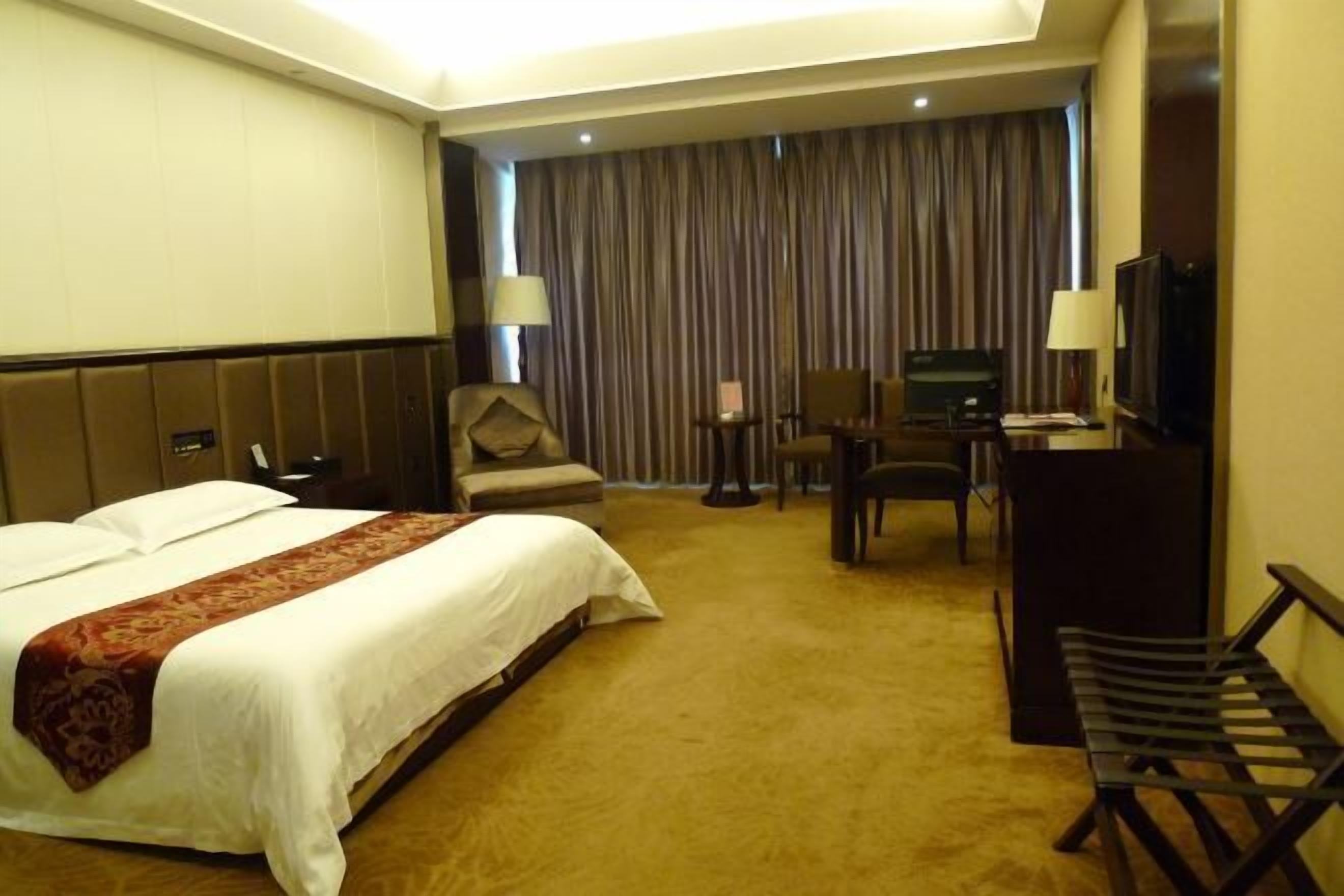 A hotel room with a large bed, a desk, a chair, a television, and a bench.