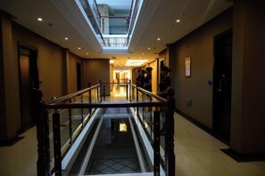 Pasillo de hotel con barandilla de vidrio, un ascensor central y un tragaluz.