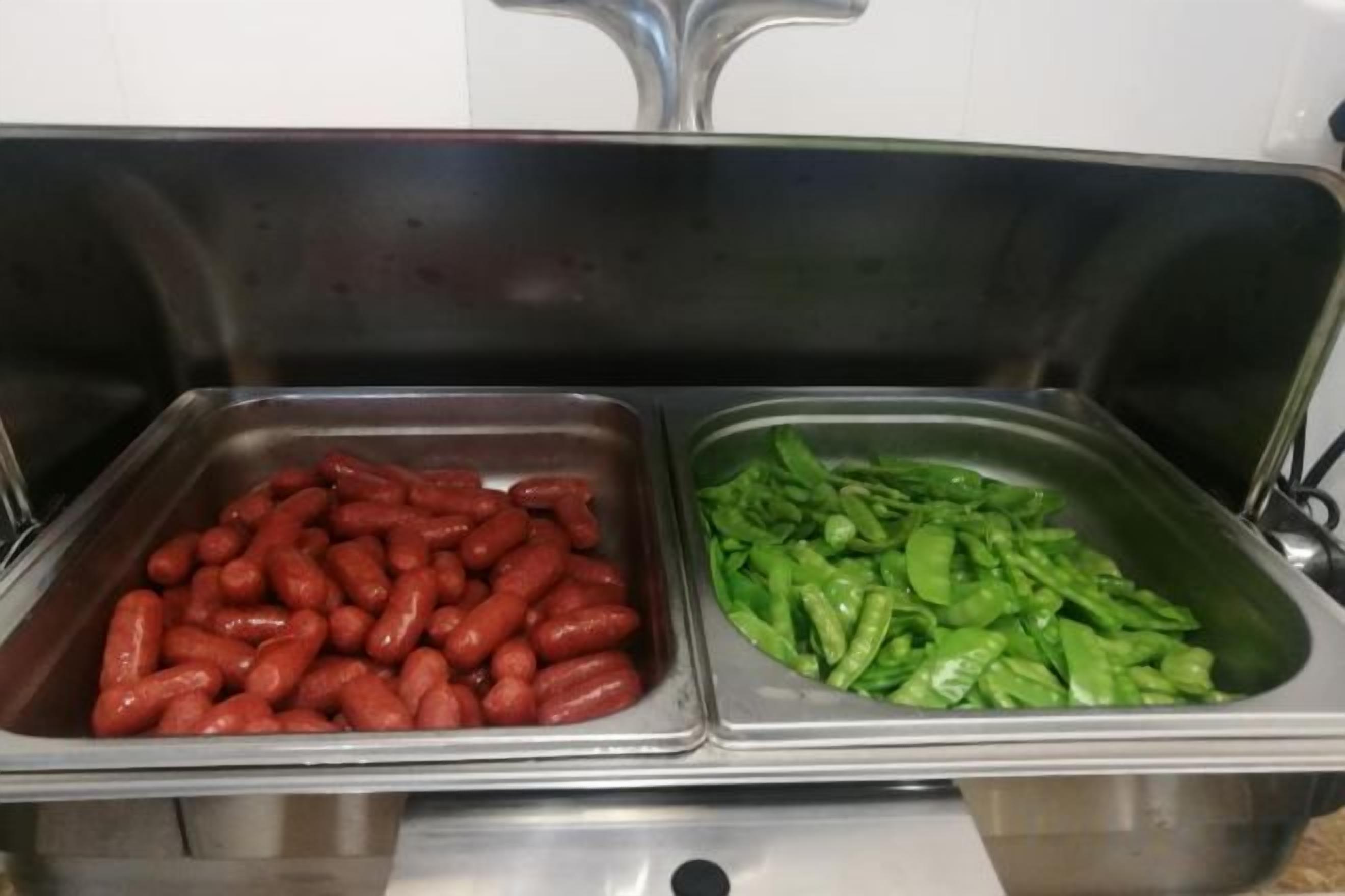 Two stainless steel food dispensers with red sausages and green vegetables.