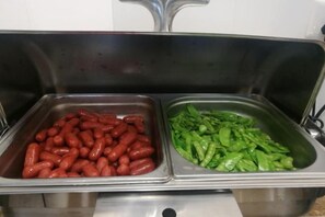 Dois dispensadores de aço inoxidável com salsichas vermelhas e vegetais verdes.