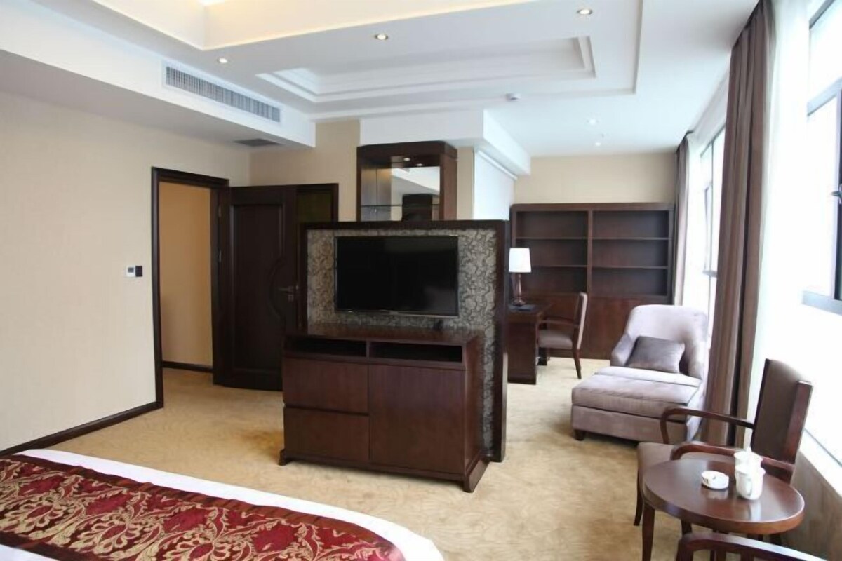 Habitación de hotel con cama, televisión sobre un mueble de madera, una silla, una mesita y una estantería.