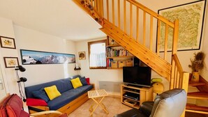 A cozy living room with a wooden staircase, a blue sofa with yellow cushions, a leather armchair, and a flat-screen TV.