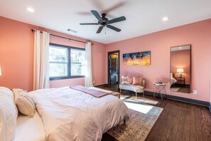 Een slaapkamer met een bed, een plafondventilator, een raam met gordijnen, een schilderij, een stoel, een klein tafeltje en een spiegel.
