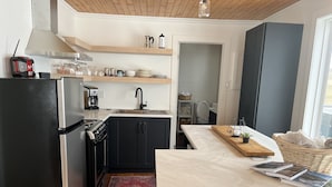Eine kompakte Küche mit einem schwarzen Kühlschrank, einem Herd und einem Spülbecken. Über dem Spülbecken befinden sich Regale mit verschiedenen Küchenutensilien. Ein Holzboden und ein roter Teppich sind zu sehen.