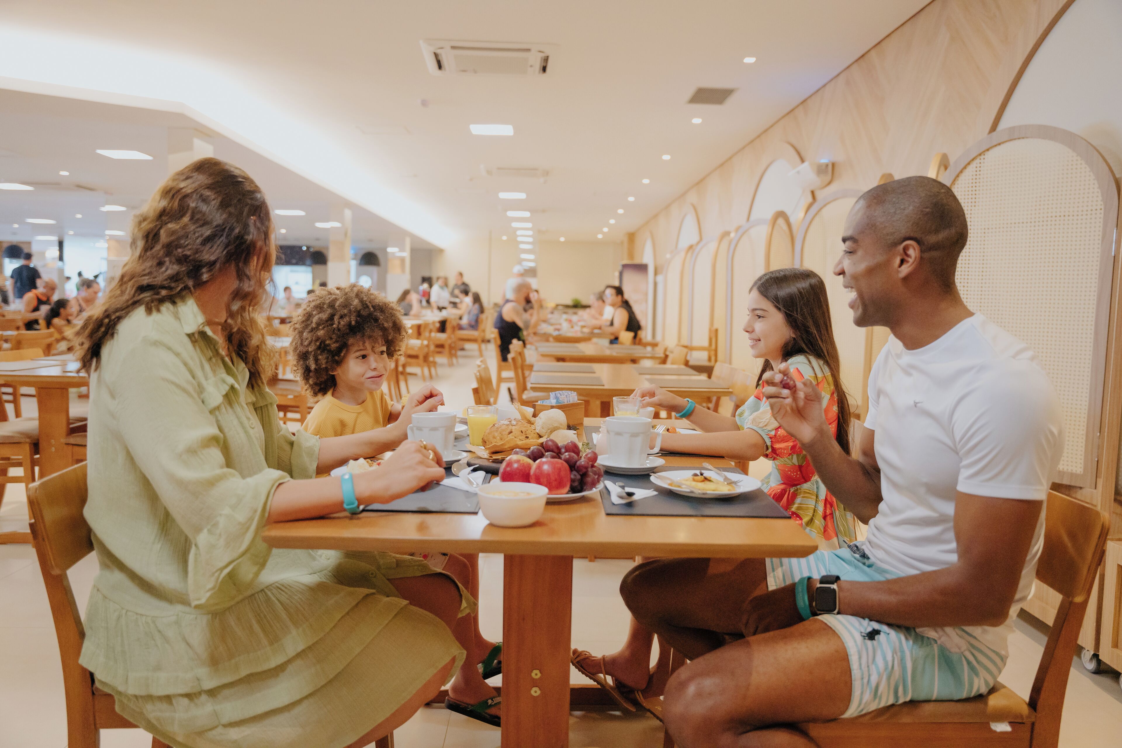 A family dining in a cafeteria with wooden tables and chairs, a variety of food items on the tables, and other diners in the background.