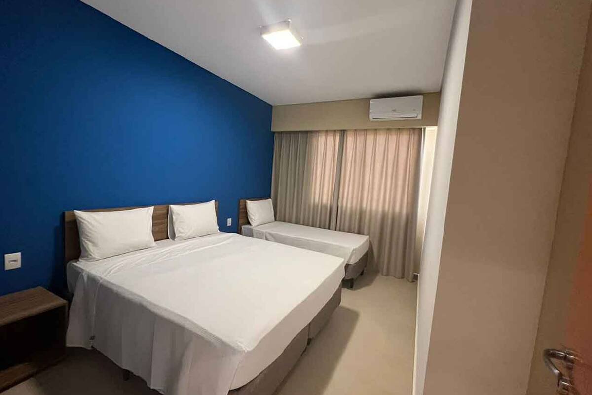 Une chambre d’hôtel avec un grand lit, deux petits lits, un mur bleu, une fenêtre avec des rideaux et un appareil de climatisation.