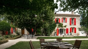 A stone building with red shutters, a dining table with chairs, and a garden path.
