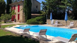 A swimming pool with lounge chairs and umbrellas, surrounded by greenery and a stone building with red shutters.