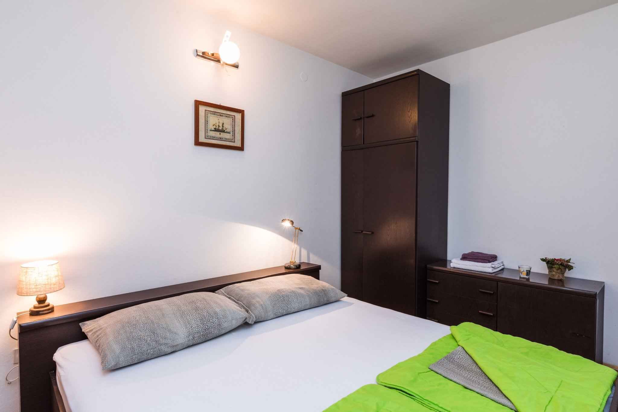 Ein ordentlich bezogenes Bett mit Kissen, eine Nachttischlampe, ein gerahmtes Bild an der Wand und ein dunkler Holzschrank.