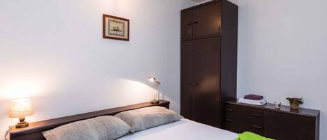 Ein ordentlich bezogenes Bett mit Kissen, eine Nachttischlampe, ein gerahmtes Bild an der Wand und ein dunkler Holzschrank.