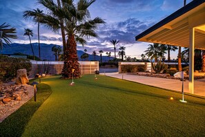 Un parcours de golf en gazon synthétique, bien entretenu, avec un club-house moderne, des palmiers et des montagnes en arrière-plan.