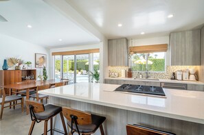 Une cuisine moderne dotée d’un grand îlot central, de chaises en bois et d’un coin repas offrant une vue sur l’extérieur.