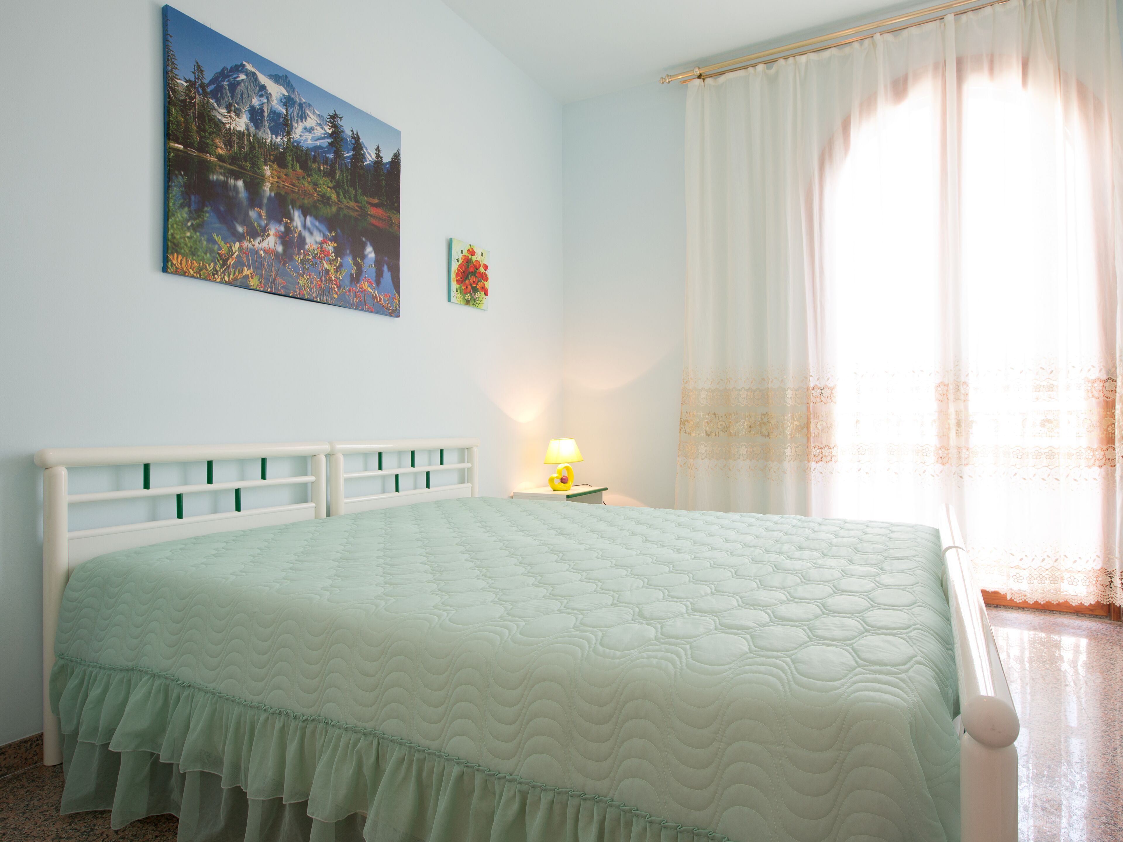 A bedroom with a bed, a painting, a lamp, and a window with curtains.