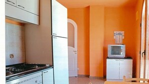 Une cuisine compacte avec des meubles blancs, une cuisinière et un petit réfrigérateur. On y trouve une table à manger avec des chaises et un pot décoratif posé dessus.