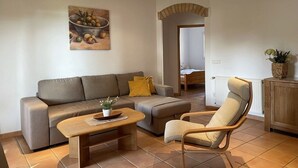 Ein Wohnzimmer mit einer Couch, einem Holztisch und einem Sessel. An der Wand hängt ein Gemälde und auf einer Kommode steht eine Pflanze.