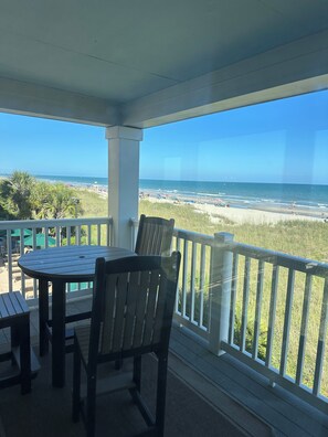 A covered outdoor seating area with a table and chairs, overlooking a beach with people and umbrellas. - Coastal Vista Charm + Seaside Paradise Retreat (North Myrtle Beach)