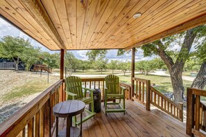 A wooden deck with two Adirondack chairs and a small round table, overlooking a grassy area with trees and a distant building.