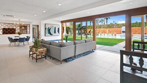 Un salon moderne avec un canapé d’angle gris, un coin repas avec des chaises bleues et une grande fenêtre donnant sur une piscine et un jardin.