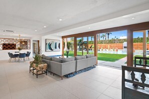 Un soggiorno moderno con un divano angolare grigio, un'area pranzo con sedie blu e un'ampia finestra che si affaccia su piscina e giardino.