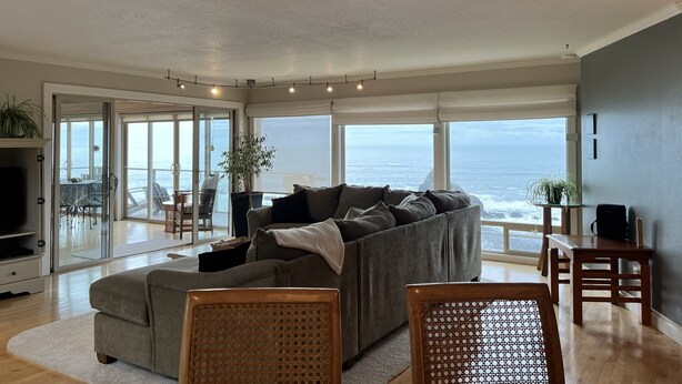 A spacious living room with a grey sectional sofa, a wooden coffee table, and a dining table with chairs. Large windows offer a view of the ocean.