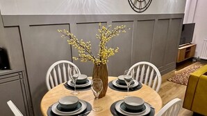 A dining area with a round table set for four, surrounded by white chairs. There is a vase with yellow flowers on the table, and a wall clock above it. - Townhouse near to Castle, Cathedral and Bailgate (Lincoln)