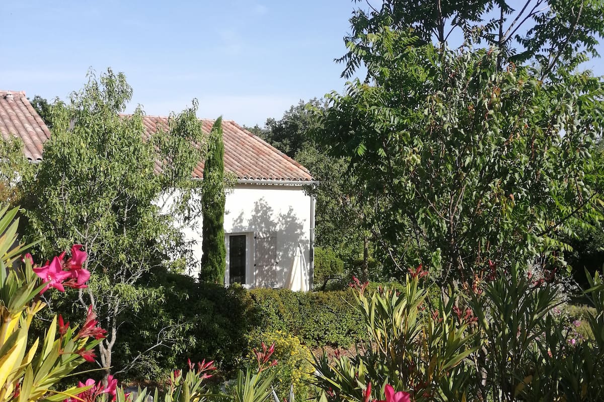 Un edificio blanco con techo de tejas, rodeado de abundante follaje y plantas con flores.