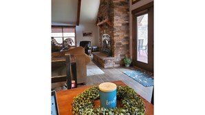 A cozy living room with a stone fireplace, a wooden table with a candle, and a view of the outdoors through large windows. - Cottage Bliss Mountain Oasis (Rexford)