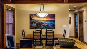 A dining area with a wooden table, four chairs, a large painting of a sunset over the ocean, and a hanging light fixture. - Cottage Bliss Mountain Oasis (Rexford)