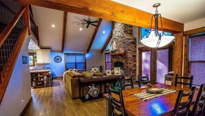 A spacious dining area with a wooden table and chairs, a stone fireplace, and a view of the kitchen in the background. - Cottage Bliss Mountain Oasis (Rexford)