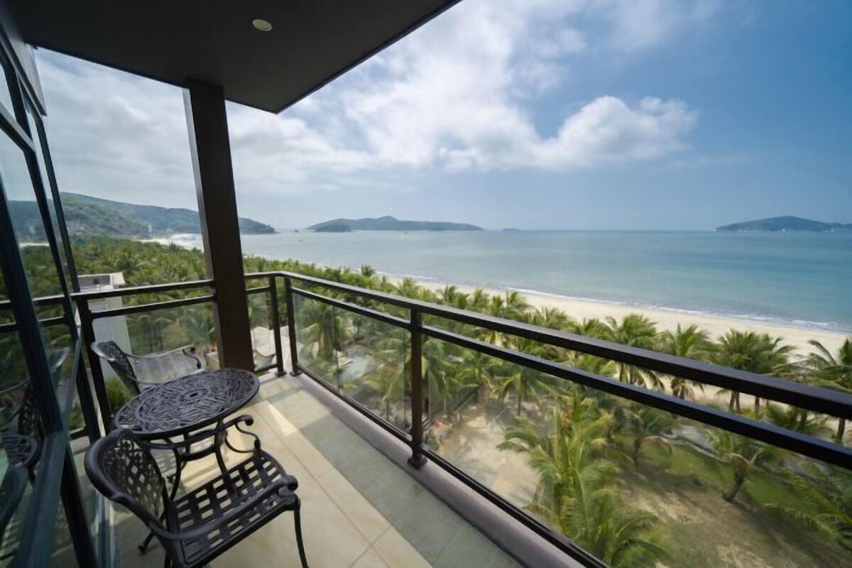 A balcony with a table and chairs overlooking a beach with palm trees and distant mountains.